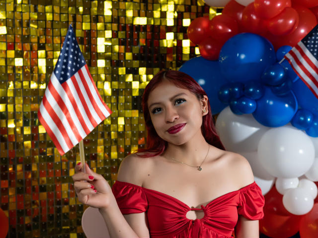 ZoeRedds, met rood haar, houdt een kleine Amerikaanse vlag vast, draagt een rode jurk met een open schouder, glimlacht en staat voor ballonnen in de kleuren rood, wit en blauw..