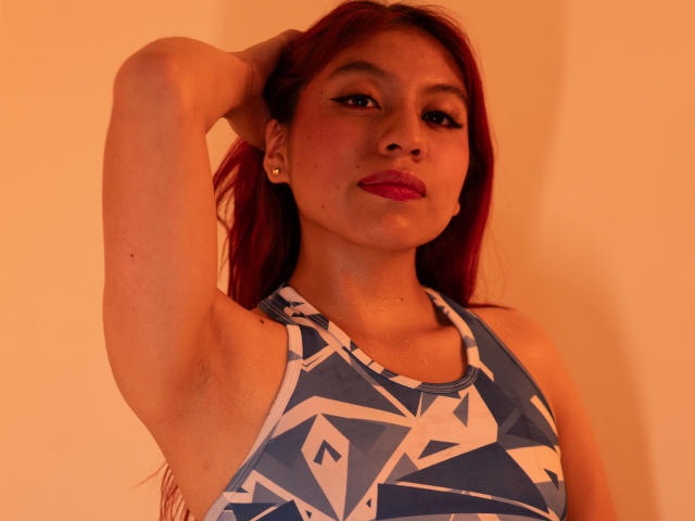 ZoeRedds with red hair, wearing a blue geometric pattern tank top, arm raised, looking at camera, red lipstick, subtle makeup, warm light, small earrings, confident expression.