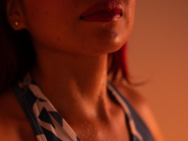 Los labios rojos y brillantes de ZoeRedds, una pequeña peca y la piel luminosa, se ven en una blusa con un estampado azul y blanco, con una iluminación naranja cálida. Su cabello rojo se ve parcialmente..