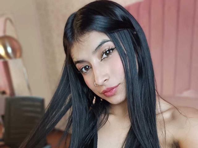 NaiaSanders with long black hair, nose ring, and subtle makeup, looking at the camera with a slight smile, wearing a black top, showing her bare shoulders.