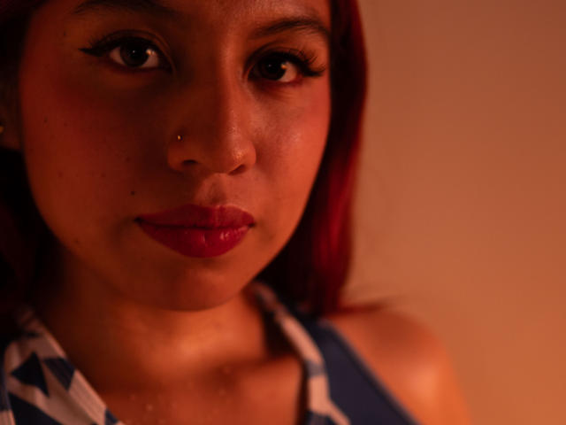 ZoeRedds ha il rossetto rosso, l'eyeliner scuro, un piccolo piercing al naso e indossa una maglietta a righe blu e bianche. La sua pelle ha una tonalità calda e ha i capelli rossi..