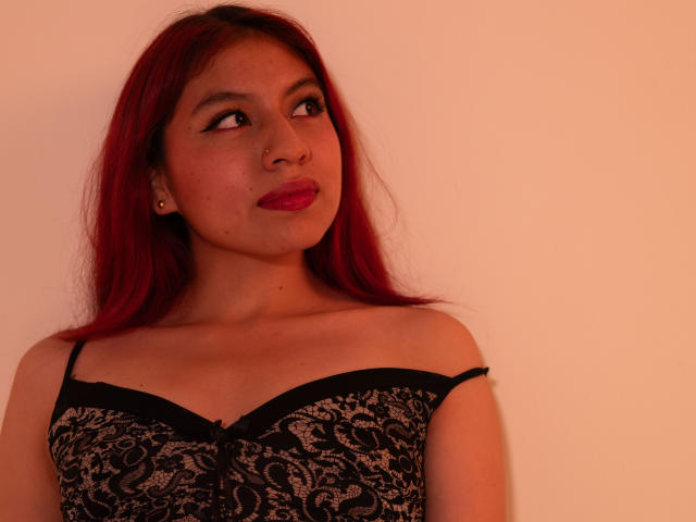 ZoeRedds with red hair, wearing a black lace dress, has small tits, a nose ring, and red lipstick, looking to the side with a slight smile. Warm light.