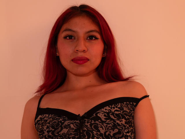 ZoeRedds has red hair, medium brown skin, and wears a black lace bra with thin straps. Her boobs are visible, and she's looking at the camera with red lipstick.