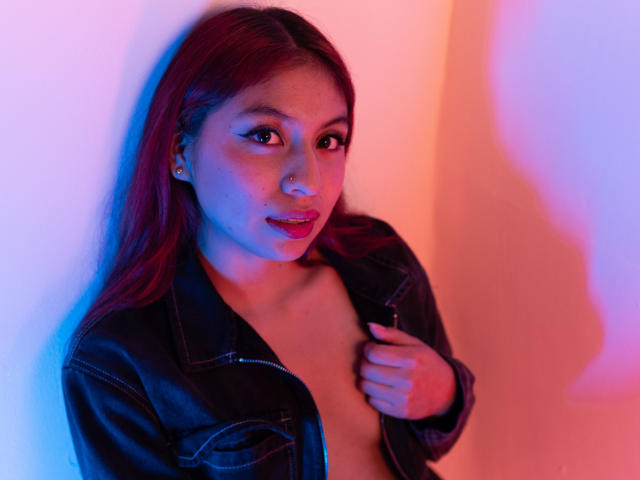 ZoeRedds with red hair, open black leather jacket, and exposed chest, holding jacket, blue and pink lighting, looking at camera with slightly parted red lips.
