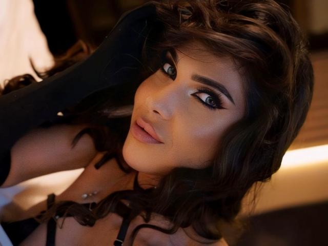LovelyKinsley tiene el pelo castaño ondulado, cejas oscuras y gruesas, y un piercing en la nariz. Lleva ropa interior negra, con un piercing en los pezones visible. Sus ojos son azules y está mirando a la cámara..