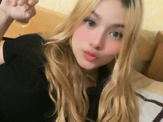 Shantal, con lunghi capelli biondi, guance rosate e labbra piene, indossa una maglietta nera, guarda la fotocamera con un leggero sorriso e alza un dito..