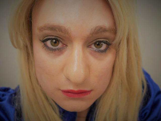 O rosto de ChantalCross-hot, cabelo loiro, olhos castanhos, batom vermelho, camisa azul, olhando para baixo com uma expressão séria, maquiagem discreta nos olhos, lábios ligeiramente entreabertos..