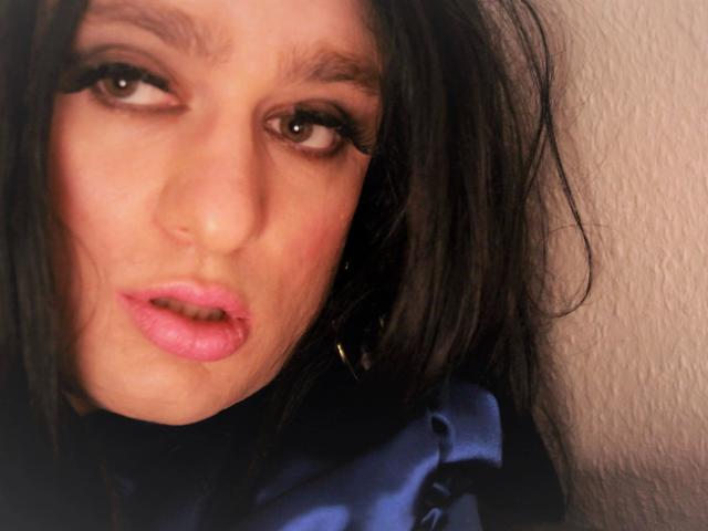 ChantalCross-hot has pink lipstick, dark eyeliner, and slightly parted pink lips. Her brown eyes are looking to the side, with messy black hair and a blue satin shirt.