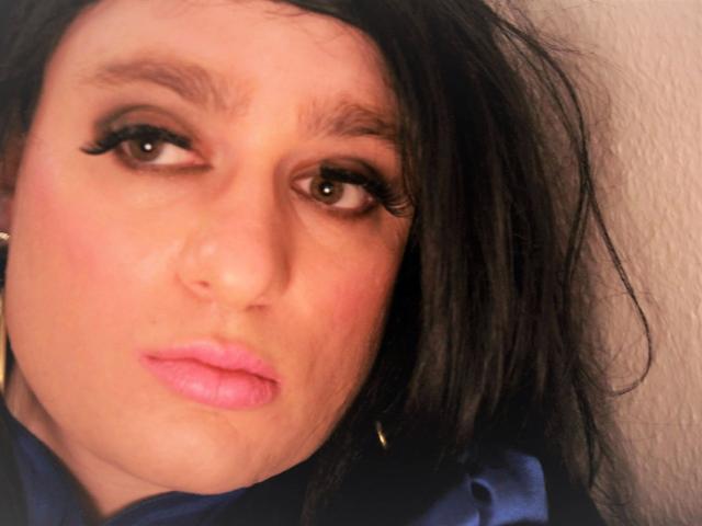 ChantalCross-hot tem lábios rosados e volumosos, delineador escuro e sobrancelhas pretas e grossas. Seu cabelo preto está desalinhado, e ela está vestindo uma camisa azul com brincos dourados..