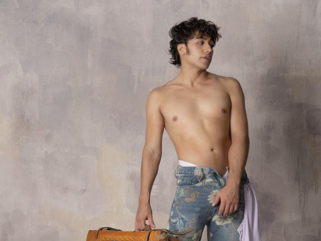 TonyDan is shirtless with a toned, muscular build, wearing unbuttoned blue jeans showing white underwear, standing next to a brown leather bag, looking to the side.
