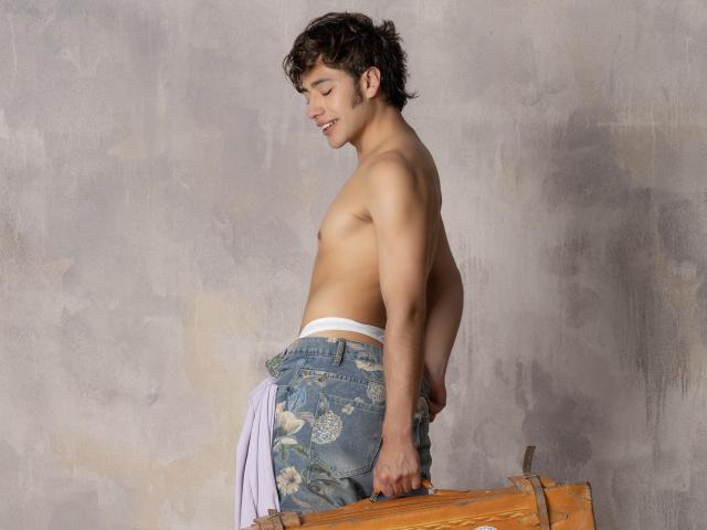 TonyDan sem camisa, com cabelo castanho encaracolado, sorrindo, vestindo calças jeans com estampas florais, roupa íntima branca, segurando uma tábua de madeira, em perfil lateral, segurando uma camisa rosa na mão direita..