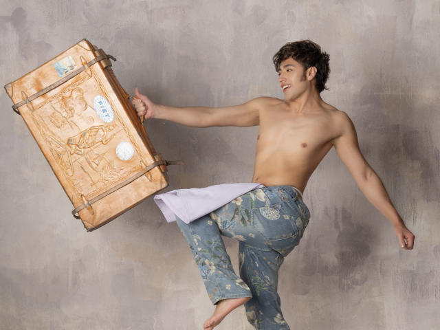 TonyDan, torse nu, avec des fesses fines et musclées, porte un pantalon bleu à motifs floraux et pointe du doigt un coffre en bois orné avec une balle blanche dessus, tout en souriant et en étant en plein saut..