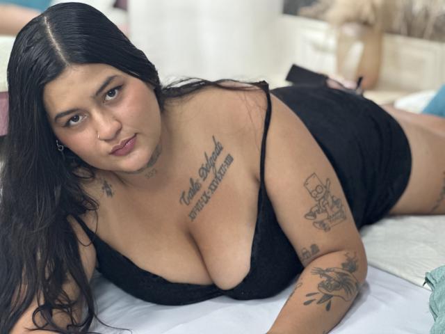 LilyRocks with long black hair, medium tits, tattoos on chest and arms, lying on bed in black lace lingerie, looking at camera with a serious expression.