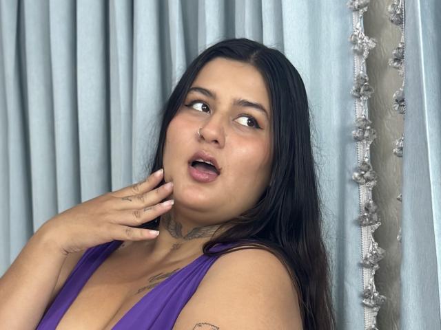LilyRocks has long black hair, a nose ring, and tattoos on her neck and hand. She's wearing a purple dress, looking surprised with her mouth open. Her hand touches her chin.