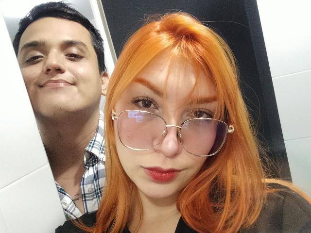 Loren has bright orange hair, glasses, and red lipstick. Ethan69 stands behind her, smiling. He's wearing a plaid shirt. Loren's tits are small, Ethan69's face is slightly blurry.