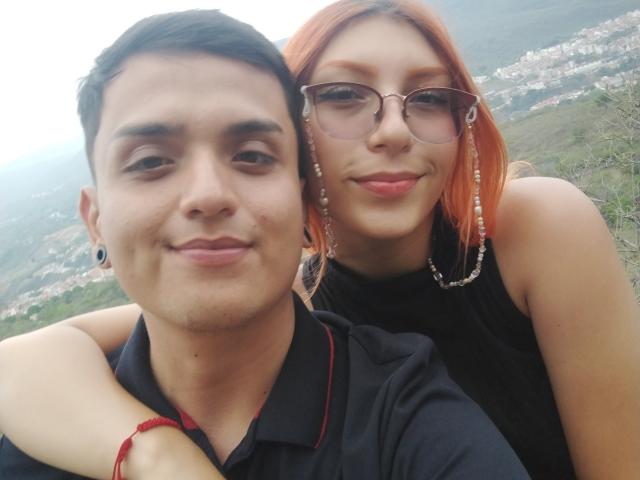 Loren has red hair, glasses, and a black sleeveless top with earrings. Ethan69 has short black hair, a black polo, and a red bracelet. Loren's arm is around Ethan69's shoulder.