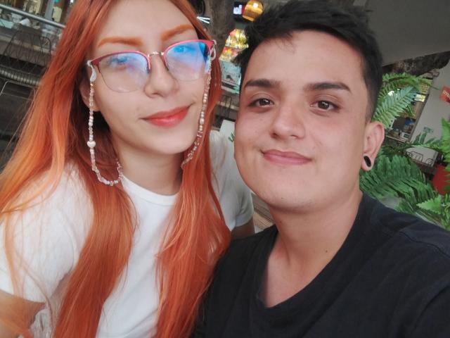 Loren with long red hair, glasses, and white shirt, next to Ethan69 with short black hair, black shirt, and ear gauges, both smiling at the camera.
