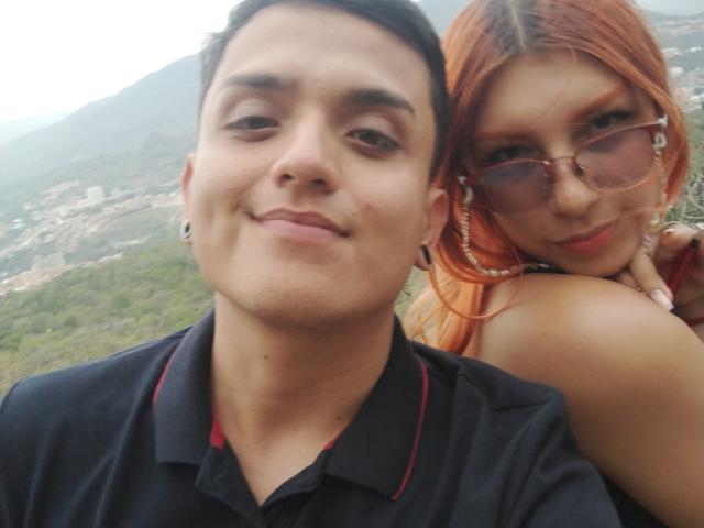 Loren has red hair, glasses, and is leaning on Ethan69's shoulder. Ethan69 has short black hair, a black polo, and a slight smile. Both are looking at the camera.