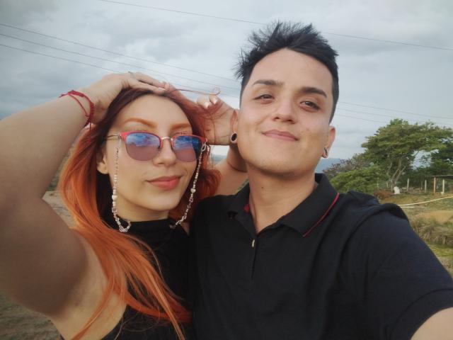 Loren has red hair, blue aviator sunglasses, and a bracelet, holding Ethan's head. Ethan has black spiky hair, black polo, and ear gauges, smiling. Outdoor selfie.