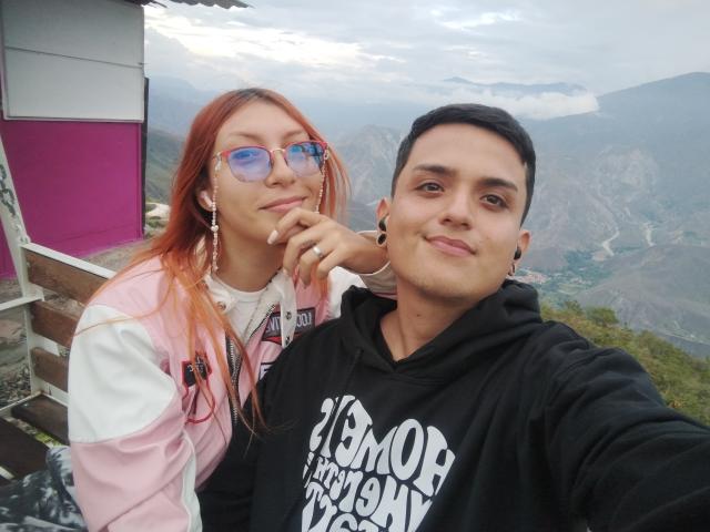 LorenandEthan69 with red hair, glasses, and pink jacket, next to Ethan69 with black hair, ear gauges, and black hoodie, both smiling at the camera. Mountain view in background.