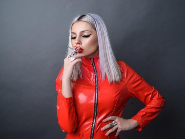 GoddessKaterina, avec ses cheveux argentés, une veste en latex rouge, fumant une cigarette, une main sur la hanche, une allure sexy et aguicheuse, un trait d'eye-liner ailé, du rouge à lèvres rouge, une bague au doigt, une expression confiante et séduisante..