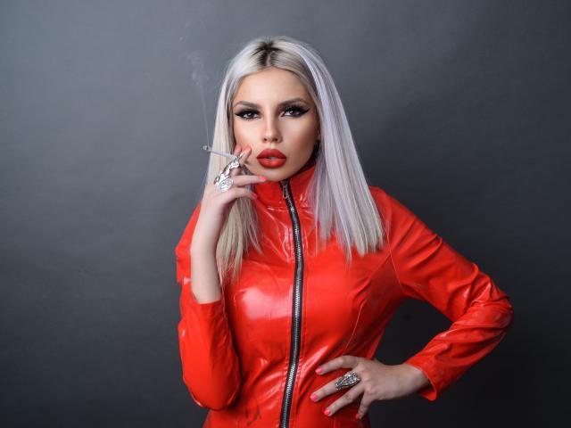 GoddessKaterina with platinum blonde hair, red latex zip-up, smoking, wearing red lipstick, ring on hand, hand on hip, looking at camera, gray background.
