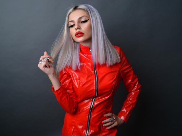 GoddessKaterina met platinablond haar, een rode vinyl jas met rits, rode lippenstift, hand op de heup, een ring vasthoudend, verleidelijk kijkend, donkere achtergrond..