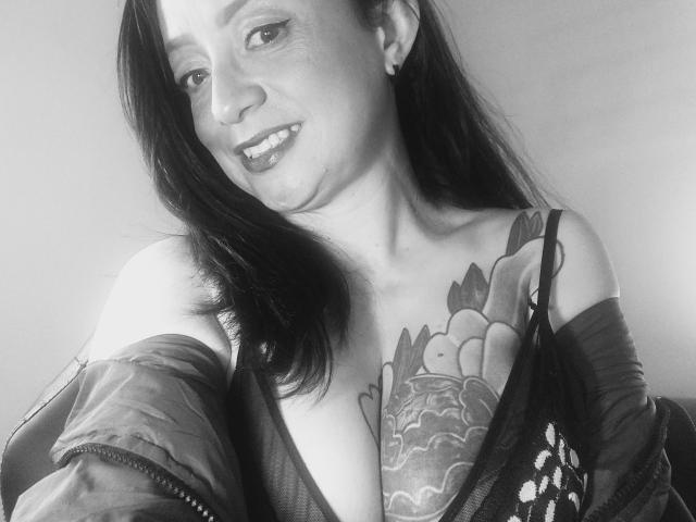 Black-and-white photo of CatalinaMonyque with long dark hair, smiling, showing large tattooed tits in a low-cut top, jacket off, earrings visible.