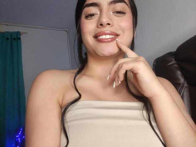 EmaRosee has a cute smile, dark hair in pigtails, white strapless top, pink lips, and is touching her chin with a finger, showing off her glossy nails.