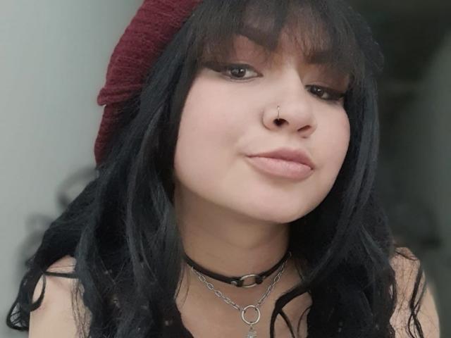 LillyMayer with black wavy hair, red beanie, black choker, and silver necklace, has a nose piercing and full pink lips, looking slightly up with a soft smile.
