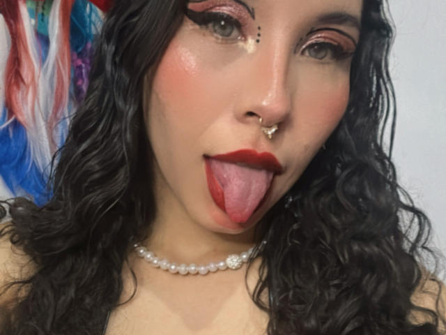 SofiBrandford with curly black hair, red lipstick, nose ring, and pearl necklace, sticking out her tongue, wearing bold pink eyeshadow, looking at the camera.