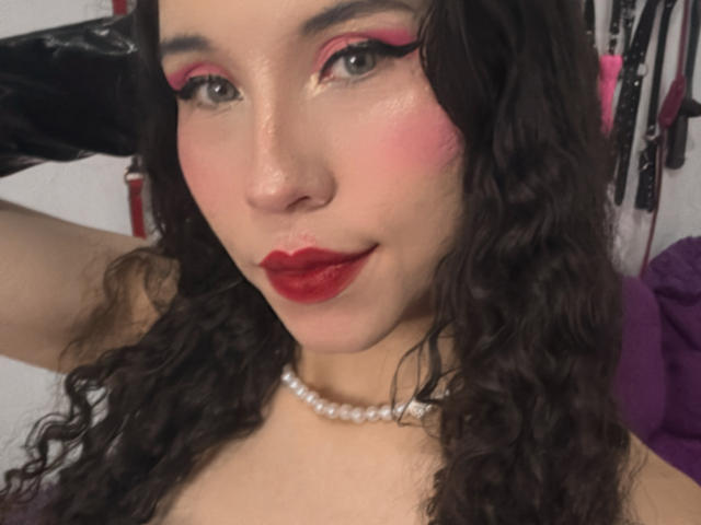 Sofi Brandford, con cabello castaño rizado, maquillaje de ojos rosa, lápiz labial rojo, un collar de perlas y un guante de látex negro en su mano derecha, mirando a la cámara con una ligera sonrisa..