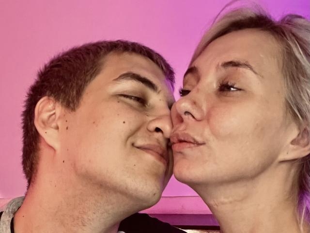 BlondIce and a dude with short brown hair and moles, noses touching, both smirking with eyes half-closed, pinkish-purple light. Dudes face to the left, BlondIce to the right.
