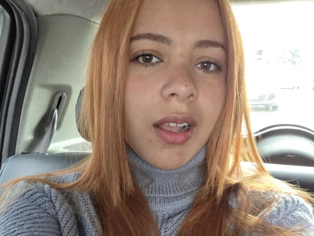 AlicePrice has light brown hair, wearing a gray turtleneck, with slightly parted pink lips, and a small beauty mark on her right cheek, sitting in a car.