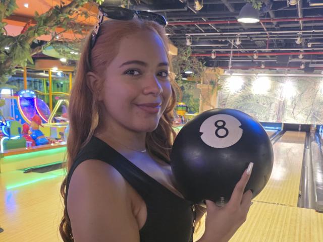 AlicePrice with red hair, black tank top, holding black bowling ball number 8, smiling, sunglasses on head, in a brightly lit bowling alley.
