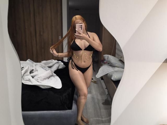 AlicePrice with long red hair, black bikini, gold chain details, holding hair, mirror selfie, phone in hand, bed with white and black covers, wooden wall in background.