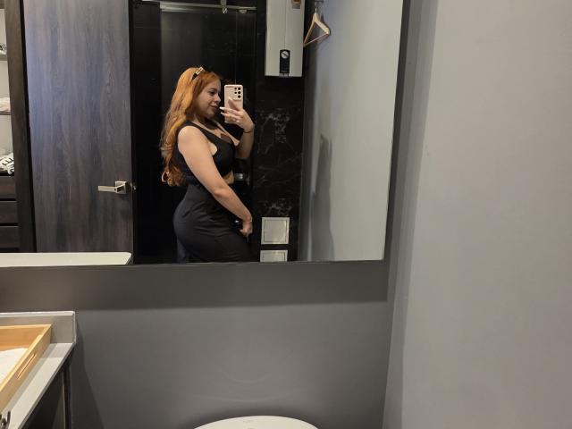 AlicePrice with long red hair, wearing a tight black dress, showing her ass in a mirror selfie, holding a pink phone, standing in a modern bathroom.