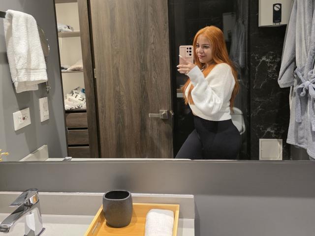 AlicePrice with long red hair, white sweater, and black pants, holding a phone, showing her curvy ass and small tits in a bathroom mirror selfie.