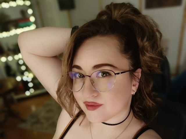 LuxNoir with glasses, brown wavy hair in a ponytail, fair skin, red lipstick, black choker, hand in hair, smiling, visible nose ring, and small earrings.
