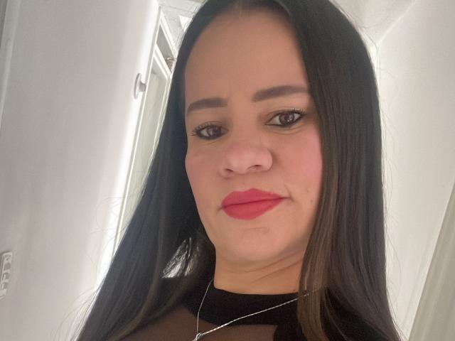 CharllotI has long black hair, red lipstick, and a slight smile. She's wearing a black sheer top and a silver necklace. Her eyebrows are thick and she has dark eyes.