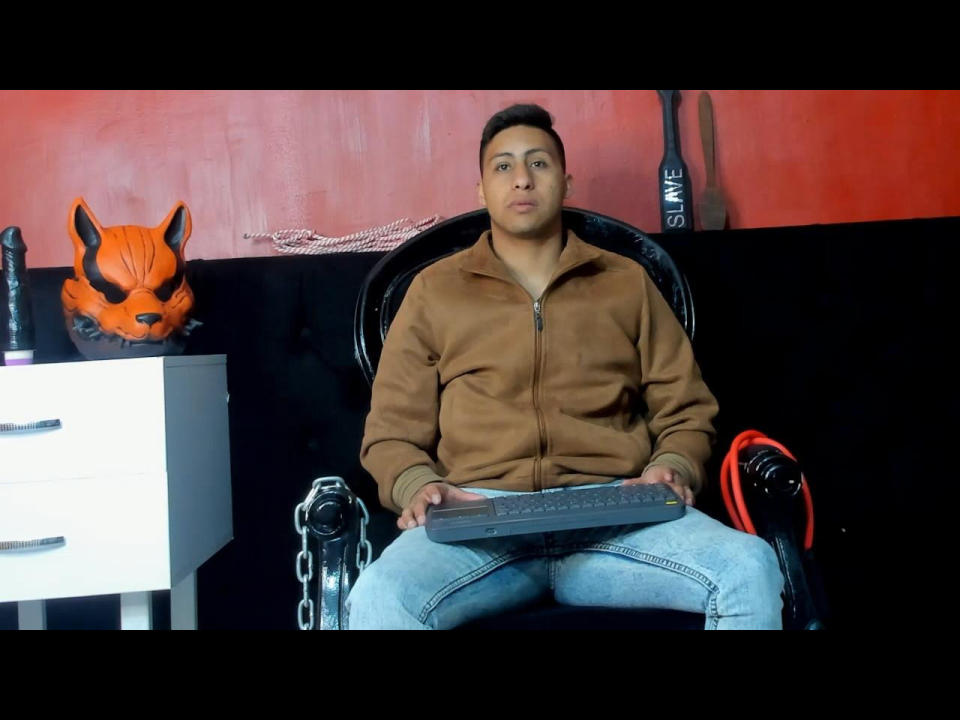 LeeoMax, chubby with short brown hair, wearing a brown zip-up jacket and jeans, sits on a black chair, holding a keyboard, with a fox mask and dildo on the left.