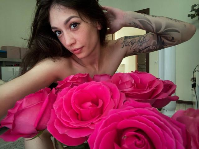 MoniFoxy mit rosa Rosen, nackt, Tattoos am rechten Arm, braunes Haar, Hand im Haar, lächelnd, rosa Rosen bedecken die Brüste, grüne Augen, helle Haut, in einem Raum..