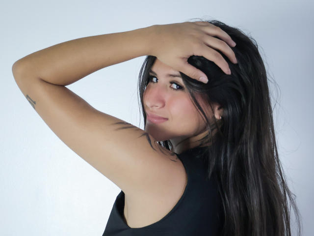 PeytonGrayy, avec de longs cheveux noirs, une peau mate, un petit tatouage sur le bras, portant un haut noir sans manches, une main dans les cheveux, un léger sourire, regardant par-dessus son épaule..