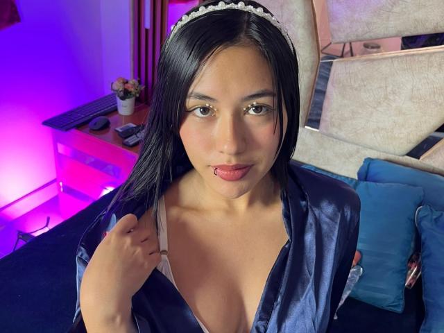 SaraHottiestX with long black hair, wearing a blue satin robe, sticking out her pink tongue, holding a white strap, looking at the camera, wearing a white headpiece.