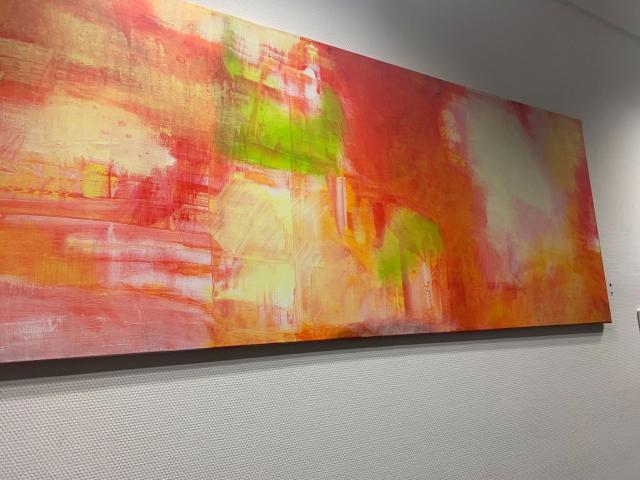A colorful abstract painting with red, orange, yellow, and green brushstrokes on a rectangular canvas, hanging on a white wall. CatwalkStar-hot's ass is in the background.