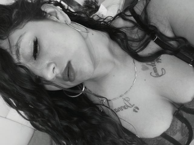 NicoleMartins with wavy black hair, hoop earrings, and a necklace, pouting her lips, showing her large tits and tattooed chest. Black-and-white selfie, lying down, looking at camera.