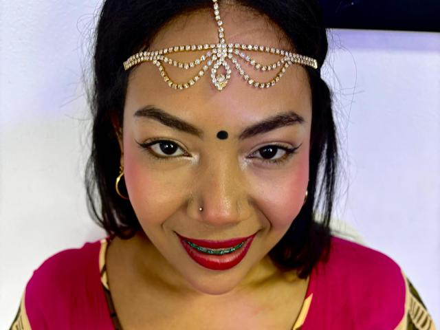 KalindaSajanii has dark brown skin, black hair, red lipstick, and a nose piercing. She's wearing a jeweled headpiece, a red and gold top, and gold hoop earrings.