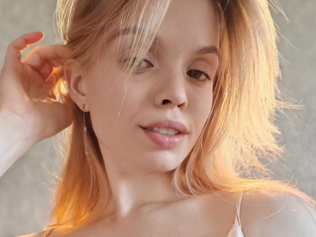 Sinteas tem cabelo loiro, pele clara e um sorriso adorável com lábios rosados e brilhantes. Ela está tocando o cabelo com a mão, mostrando uma pequena perfuração na orelha..