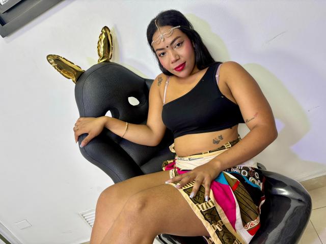 KalindaSajanii with tan skin, black hair, and red lipstick sits in a black chair with gold ears, wearing a black crop top and colorful skirt, showing her ass and tits.