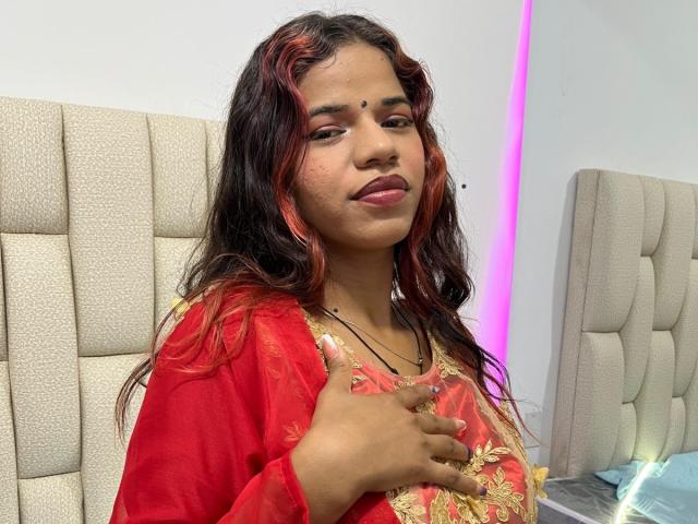 KairaDayamai with red and black wavy hair, tan skin, wearing a red floral top, holding chest, pink lipstick, nose dot, subtle smile, beige headboard, purple light.